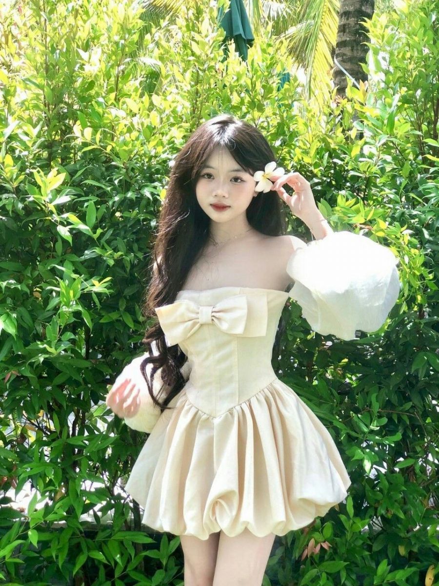 Ảnh gái trẻ mặc váy liền màu bề với tay phồng tạo dáng bên cạnh bụi cây xanh