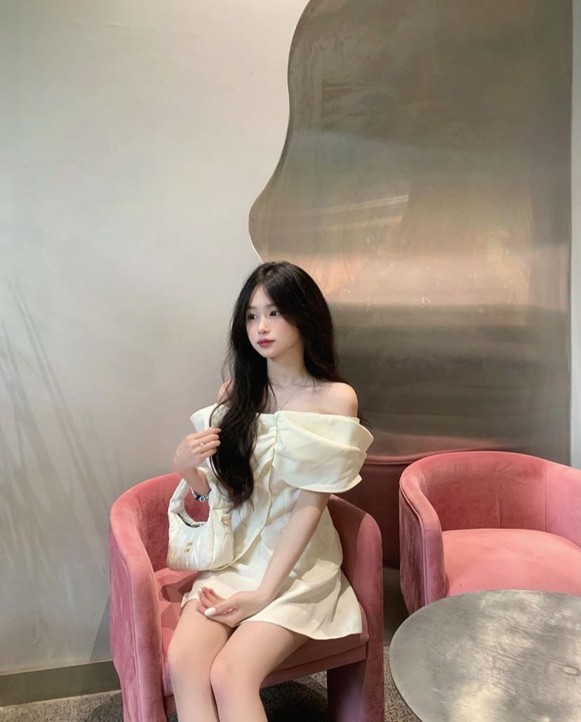 Cô gái trẻ mặc đầm trắng lệch vai ngồi trên ghế màu hồng