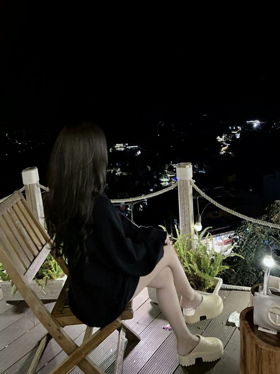 Gái xinh mặc áo sweater đen ngồi trên ghế gỗ nhìn ra thành phố về đêm