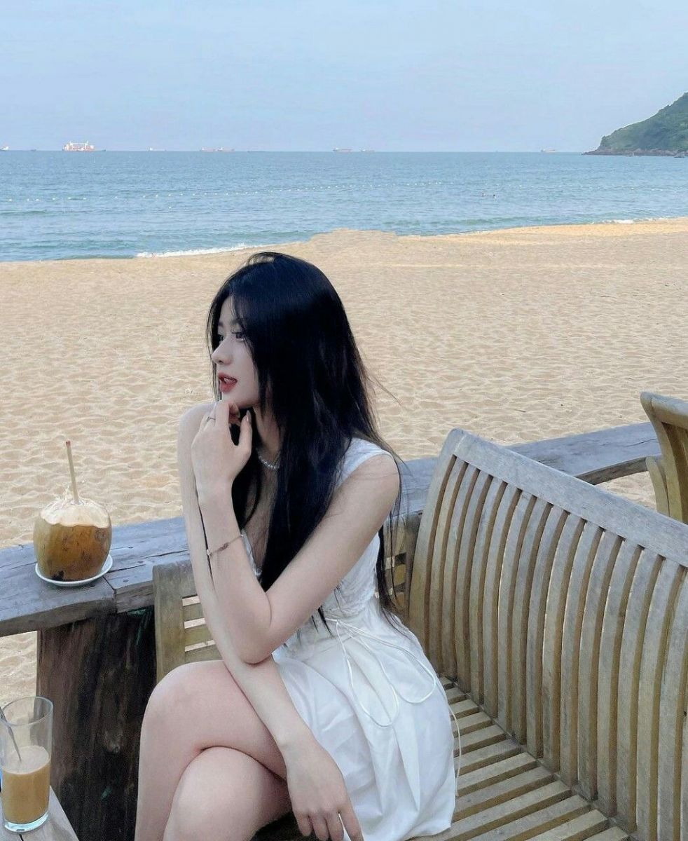 Gái xinh mặc váy trắng ngồi trên ghế gỗ bên bãi biển