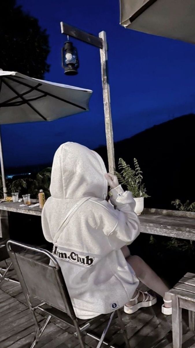 Ảnh gái trẻ mặc hoodie màu xám ngồi trên ghế tay cầm ly nước ngâm cảnh đêm tại quán cà phê