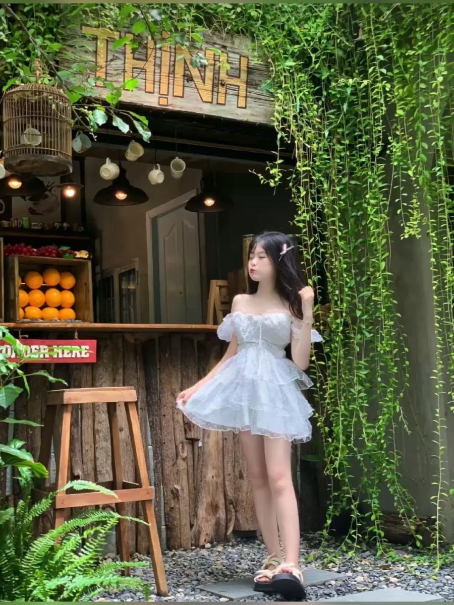 Cô gái trẻ mặc váy trắng xòe xinh đẹp trước quán cà phê xanh tươi