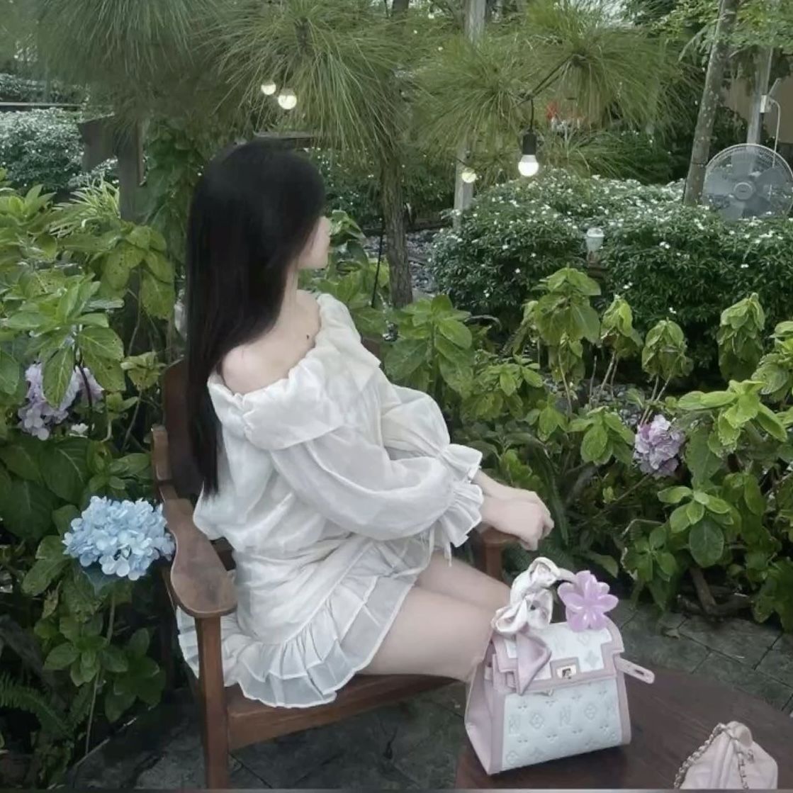 Gái xinh mặc váy trắng ngồi trên ghế gỗ giữa khu vườn xanh